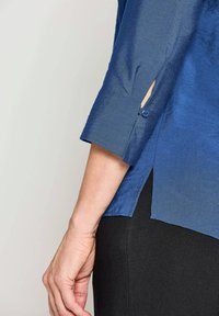 Blusa blu semi-trasparente con polsini abbottonati e spacco laterale; presenta una trama liscia e una vestibilità leggermente ampia. Abbinata a pantaloni neri.