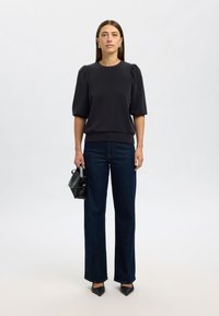 Svart topp med puffärmar och rund halsringning, kombinerad med mörkblå vida jeans. Modell som håller en liten svart handväska. Elegant presentation.