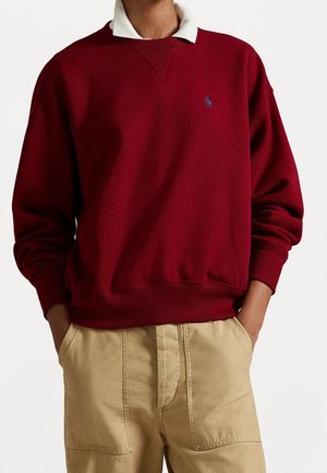 Person iført en rød sweatshirt med rund hals over en hvid skjorte med krave og lysebrune bukser, hænderne i lommerne, neutral baggrund.