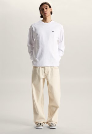 Young person standing wearing a white Vans long-sleeve shirt, loose beige pants, and black-and-white checkered slip-on shoes.