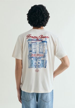 Persona con cabello rizado que lleva una camiseta blanca con una ilustración de una cafetería en azul y rojo y texto en la parte trasera, combinada con jeans azul claro.