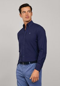 Marineblauwe knoopshirt met een puntkraag, lange mouwen en een klein logo op de borst. Beschikt over contrasterende knopen en een gladde textuur.