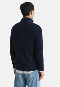 Navy-blauer Kabelstrick-Pullover mit hohem Kragen und gerippten Bündchen. Verfügt über ein strukturiertes Muster und eine lässige Passform, kombiniert mit hellblauen Jeans.