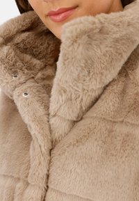 Abrigo de piel sintética beige con una textura suave, que cuenta con un cuello alto y cierres de snap plateados en el frente. Tejido estampado para mayor profundidad.