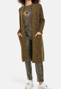 Langer, olivgrüner Cardigan mit strukturiertem, glitzerndem Strick, seitlichen Taschen. Kombiniert mit einem braunen T-Shirt mit Grafik und enganliegenden, glänzenden grünen Hosen. Weiße Sneakers.