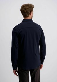 Man met lichtbruin haar, gekleed in een smalle, lange mouw donkerblauwe blouse en donkere broek, staande met zijn rug naar de camera tegen een effen achtergrond.