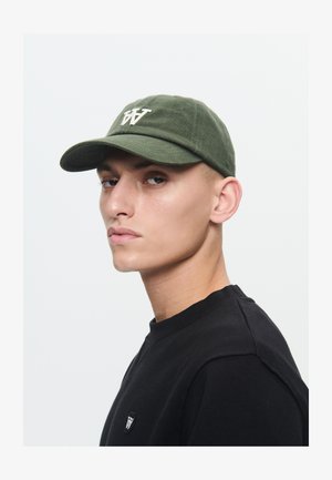 Fiatal férfi zöld baseballsapkában, melyen fehér "W" logó található, fekete pulóverben, amelyen a mellrészen egy kis, azonos "W" embléma látható.