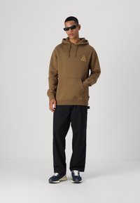 Brauner Kapuzenpullover mit einem dreieckigen gelben Logo, vorn mit Kängurutasche, kombiniert mit schwarzen lockeren Hosen und marineblauen Sneakers. Glatte Stofftextur.