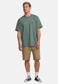 Grünes Baumwoll-T-Shirt mit Rundhalsausschnitt und kurzen Ärmeln, mit einem bedruckten Logo auf der Brust. Kombiniert mit khakifarbenen Shorts und schwarzen Sneakers.