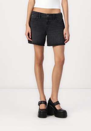 Shorts en denim noir avec un ourlet effiloché, comportant des poches avant et une fermeture à bouton. Assortis avec des chaussures à talons noires avec des brides de cheville.