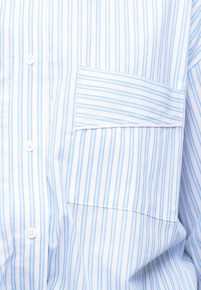 Λευκό πουκάμισο με λεπτές κάθετες μπλε ρίγες, διαθέτει μπροστινή τσέπη με οριζόντιες ρίγες και κλείσιμο με κουμπιά, σε κοντινή λήψη.