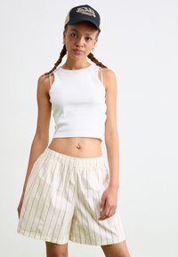 Biely rebrovaný crop top v kombinácii so svetlými smotanovými šortkami s červenými vertikálnymi pruhmi a elastickým pásom. Modelka má tiež na sebe čiernu čiapku.