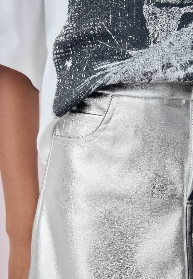 Silberne metallische Hosen mit glatter Textur und Seitentasche, kombiniert mit einem weißen Grafik-T-Shirt, das ein dunkles abstraktes Design zeigt.
