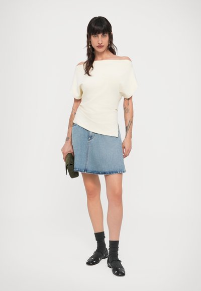 Εκρού τοπ με άνοιγμα στους ώμους και κοντά μανίκια, συνδυασμένο με μπλε τζιν φούστα. Φοριούνται μαύρα παπούτσια και μια σκούρα πράσινη τσάντα χειρός.