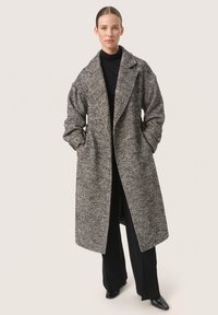Geweven zwart-wit lange jas met een ingesneden kraag, grote mouwen en voorzakken, gecombineerd met zwarte broek en schoenen.