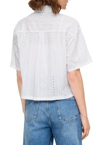 Witte T-shirt met korte mouwen en een gestructureerd ontwerp met opengewerkte patronen, een achterpand en een boxy cropped fit. G gedragen met blauwe spijkerbroek.