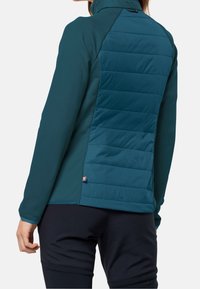 Veste bleu sarcelle avec un panneau arrière matelassé, un devant lisse, un col montant et des fentes latérales. Texture douce et design épuré.