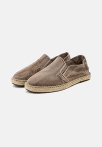 Zapatos de lona marrones tipo slip-on con paneles elásticos en los laterales, suela intermedia de cuerda de yute y superficie de tela texturizada. Punta redondeada y suela plana.
