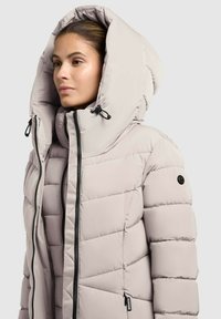 Svetlosivý pufovaný kabát s veľkou kapucňou, prešívaným dizajnom a čiernymi zipsami. Obsahuje šnúrky na stiahnutie a nášivku s logom na rukáve.