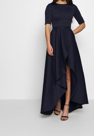 Femme portant une robe bleu marine à ourlet asymétrique, manches mi-longues aux coudes et fente avant superposée, associée à des talons noirs à bride autour de la cheville.