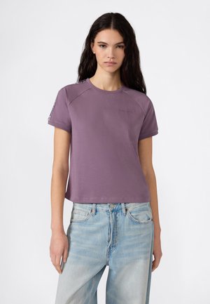 Joven con cabello largo y oscuro, vistiendo una camiseta morada de manga corta y jeans azul claro, posando sobre un fondo blanco.