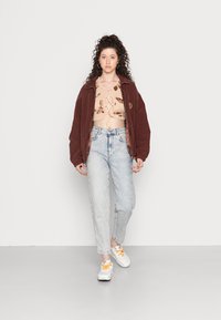Casaco oversized bordeaux, top curto de malha bege com padrões florais, jeans de cintura alta azul claro e ténis brancos com detalhes laranja.