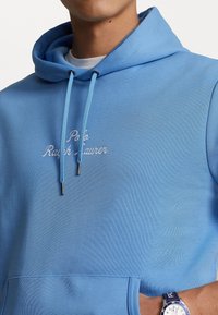 Man med ljusblå Polo Ralph Lauren hoodie med snoddar och matchande klocka på handleden, händerna i framfickan.