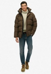 Mand iført en brun polstret jakke, en cremet rullekrave sweater, blå jeans og tan ruskindsstøvler, stående med den ene hånd i lommen.