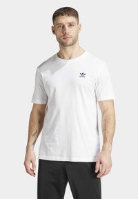 Bílý bavlněný tričko s kulatým výstřihem a krátkými rukávy, se malým černým logem Adidas na levé části hrudníku.