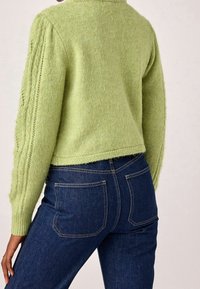 Osoba oblečená ve světle zeleném pleteném svetru s texturou a tmavě modrých denimových džínách, zobrazená zezadu s rukou odloženou na boku.