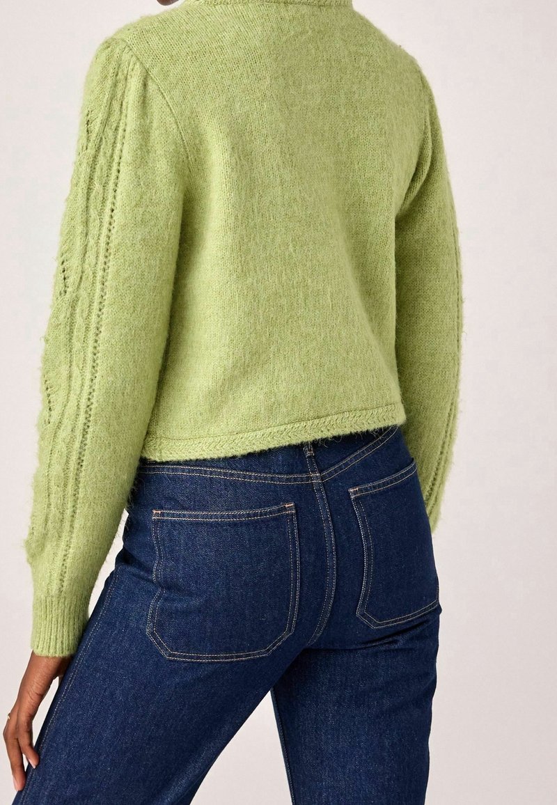 Osoba oblečená ve světle zeleném pleteném svetru s texturou a tmavě modrých denimových džínách, zobrazená zezadu s rukou odloženou na boku.