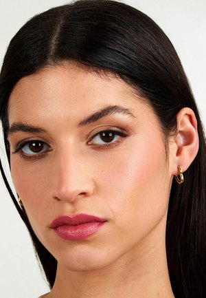 Donna con capelli scuri e lisci, che indossa orecchini a cerchio dorati, trucco naturale con ombretto marrone e rossetto rosa, rivolta in avanti con espressione neutra.