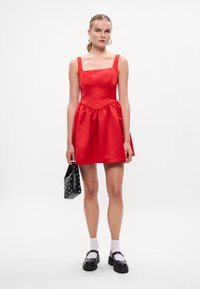 Červené saténové šaty so štvorcovým výstrihom, nariasenou sukňou a širokými ramienkami. V páre s malou čiernou taškou so cvočkami a čiernymi topánkami.