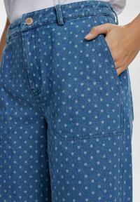 Mėlyni džinsai su taškių raštu, priekinėmis kišenėmis ir vienu mygtuku prie juosmens. Minkšta tekstūra, laisvas siluetas.