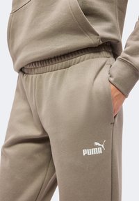 Άτομο που φοράει μπεζ φόρμα Puma και αντίστοιχο φούτερ, με το ένα χέρι στην τσέπη, δείχνοντας το λογότυπο Puma στα παντελόνια.