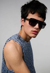 Gafas de sol rectangulares negras con lentes degradados, acentuadas con detalles de carey. Llevadas con una blusa azul sin mangas y texturizada.