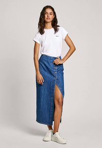 Fustă maxi din denim cu o șlițată înaltă, prevăzută cu cinci nasturi pe partea din față, asortată cu un tricou alb mulat și adidași albi.