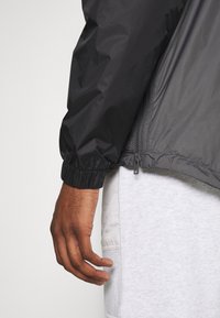 Main gauche d'une personne portant une veste noire avec un poignet élastique et un pantalon de survêtement gris clair avec une poche latérale.