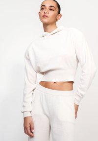 Tânără cu părul dat pe spate, îmbrăcată într-un hanorac alb scurt și o pereche de pantaloni cu talie înaltă asortați, pozând cu mâna pe șold, pe un fundal alb.