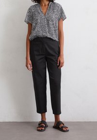 Femme portant un pantalon noir court, des sandales plates noires avec une boucle dorée, et un chemisier à manches courtes noir et blanc à motifs, debout contre un mur blanc.