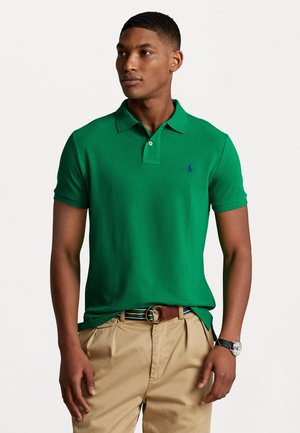 Polo vert en coton, avec un col, des manches courtes et un petit logo bleu sur la poitrine, associé à un pantalon beige et une ceinture.