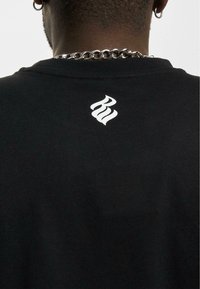 Juodas padengtas audinys marškinėliai su baltu stilizuotu logotipu gale prie kaklo. Dizainas paprastas, be jokių papildomų raštų ar detalių.