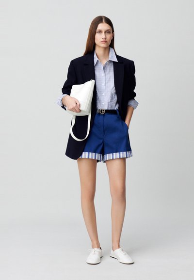 Blazer marine sur chemise à carreaux bleus, associé à un short bleu avec un bord à carreaux. Un sac et des chaussures blanches complètent le look.
