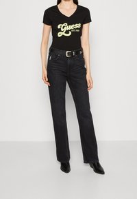 Sort bomulds t-shirt med en gul "Guess EST. 1987" grafik, parret med mørk denim flair jeans og sorte ankelstøvler.