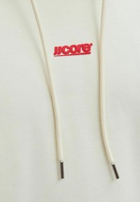 Tela de sudadera color crema con cordones blancos que terminan en puntas plateadas y pequeño logo bordado en rojo "JJCORE" encima del centro.