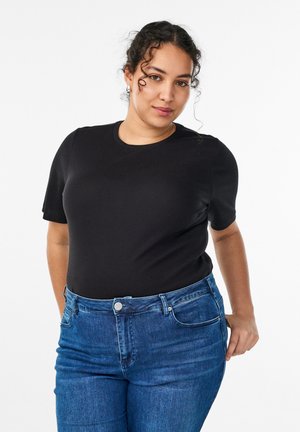 Jeune femme aux cheveux bouclés portant un haut noir à manches courtes et un jean bleu, posant avec une main dans la poche contre un fond blanc.