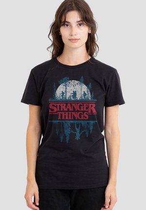 Young woman wearing black T-shirt with "Stranger Things" text and graphic of kids on bikes and upside-down figure in forest background.
