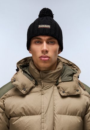 Zwarte gebreide beanie met een pompon en logo-patch, gecombineerd met een tan gekleed pufferjack met olijf-groene schouderaccenten.