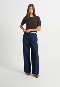 Brązowa dzianinowa bluzka z krótkim rękawem w połączeniu z wysokim stanem i szerokimi nogawkami w ciemnoniebieskim denimu. Modelka nosi beżowe buty na wysokim obcasie.