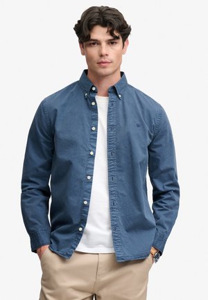 Ung mand med mørkt bølget hår iført en blå skjorte med knapper over en hvid t-shirt og beige bukser med hænderne i lommerne.